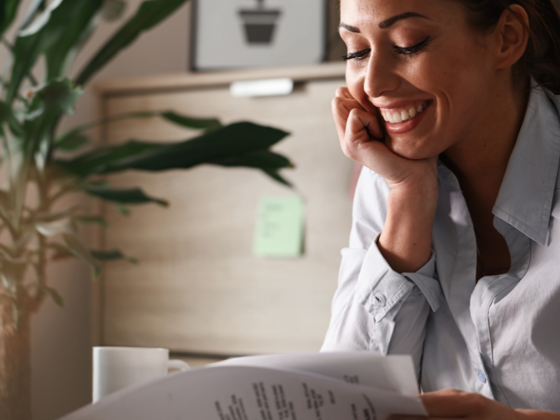 A imagem mostra uma mulher sorridente lendo documentos em um ambiente interno, provavelmente um escritório ou espaço de trabalho pessoal.