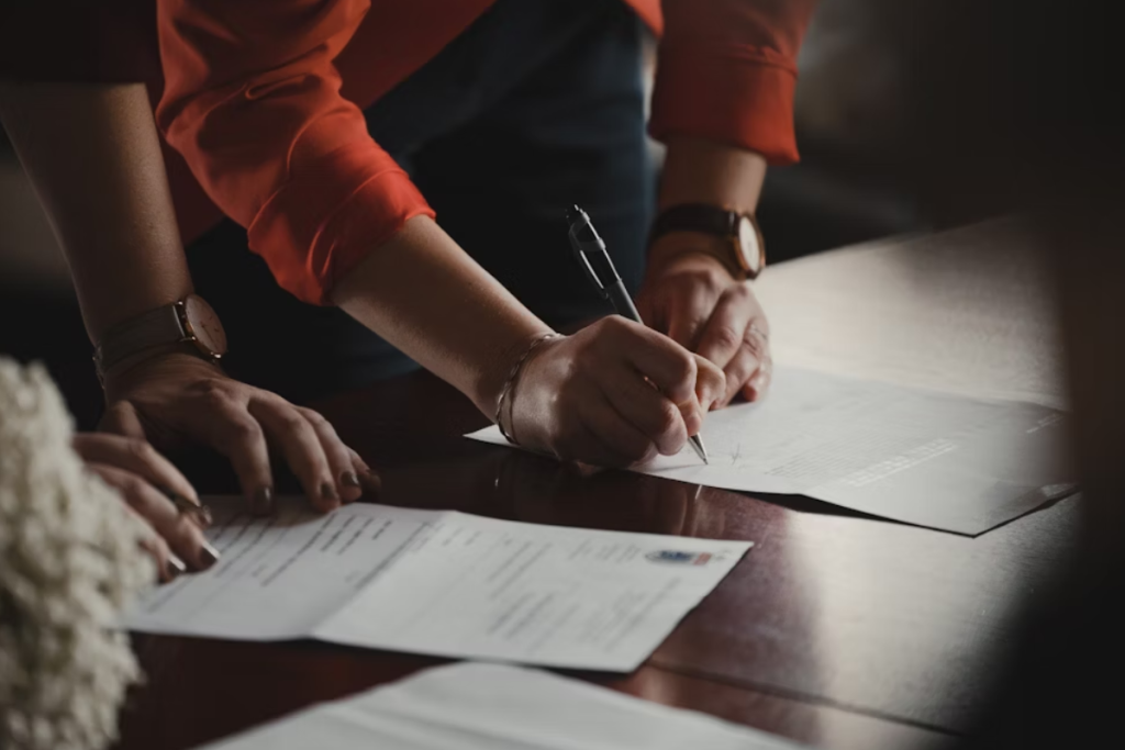 A imagem mostra uma pessoa assinando documentos sobre uma mesa, representando uma situação formal e administrativa. A cena transmite seriedade, compromisso e envolvimento com questões contratuais ou burocráticas.