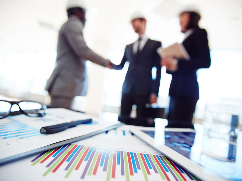 A imagem mostra um ambiente corporativo de negócios com foco em documentos e gráficos financeiros em primeiro plano, enquanto ao fundo, de forma desfocada, ocorre uma interação entre profissionais executivos.