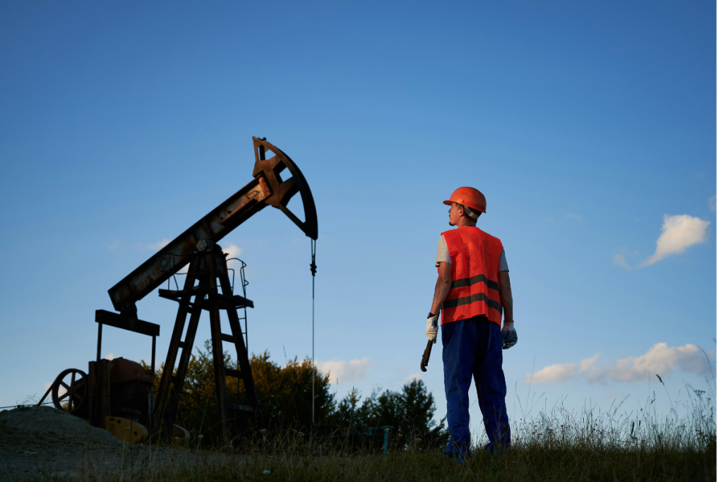 A imagem retrata um cenário típico de extração terrestre de petróleo (onshore), com um trabalhador observando uma bomba de petróleo.