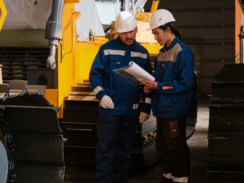 A imagem mostra dois trabalhadores industriais em um ambiente interno de manutenção ou produção, provavelmente relacionado ao setor de máquinas pesadas ou infraestrutura industrial.