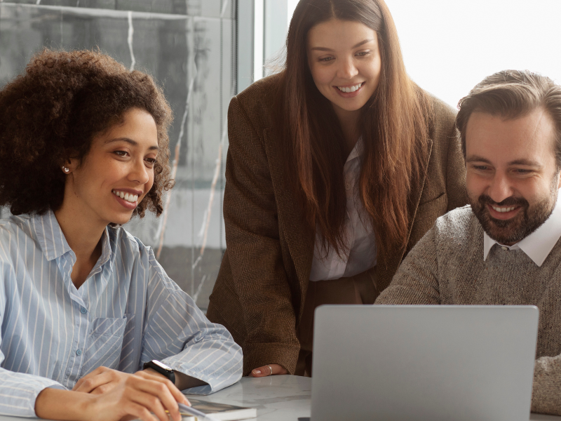 A imagem mostra três colegas de trabalho reunidos ao redor de um notebook, sorrindo e aparentemente interagindo com algo positivo ou interessante na tela. A atmosfera é descontraída, colaborativa e profissional.