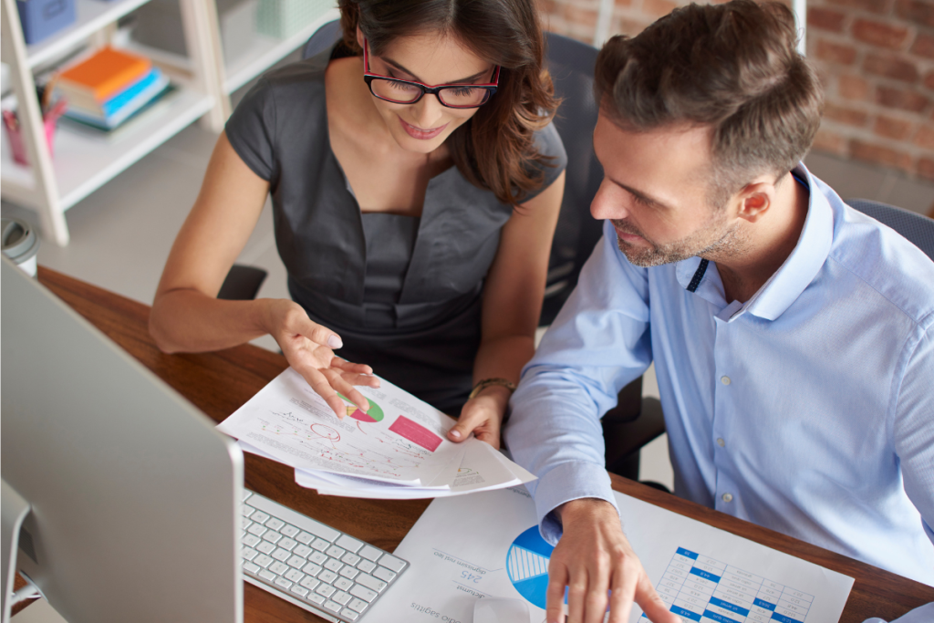 A imagem mostra dois profissionais, um homem e uma mulher, trabalhando juntos em um ambiente de escritório moderno, focados em analisar gráficos e relatórios financeiros.