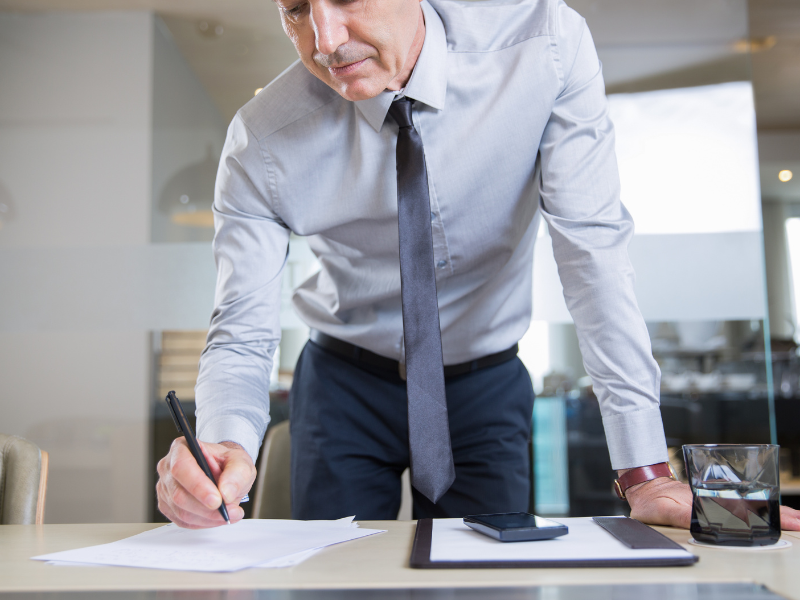 A imagem mostra um homem de negócios em um ambiente corporativo, inclinado sobre uma mesa enquanto escreve em um papel. Ele está vestido com uma camisa social cinza clara, gravata escura e calça social, transmitindo uma aparência formal e profissional.