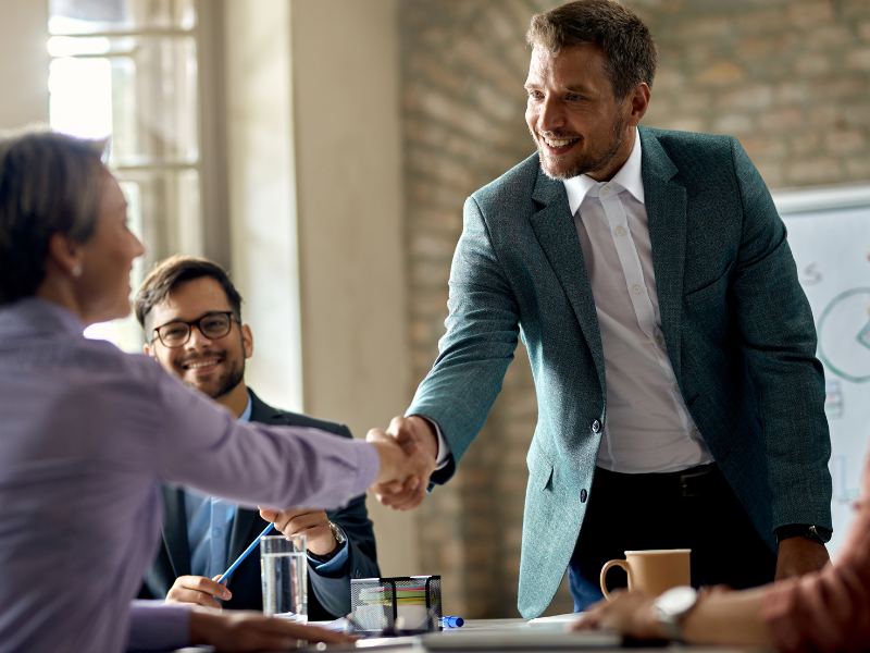 A imagem mostra um ambiente corporativo ou de reunião de negócios, com destaque para duas pessoas apertando as mãos, sinalizando fechamento de acordo, parceria ou recepção formal. A cena transmite profissionalismo, cordialidade e colaboração.