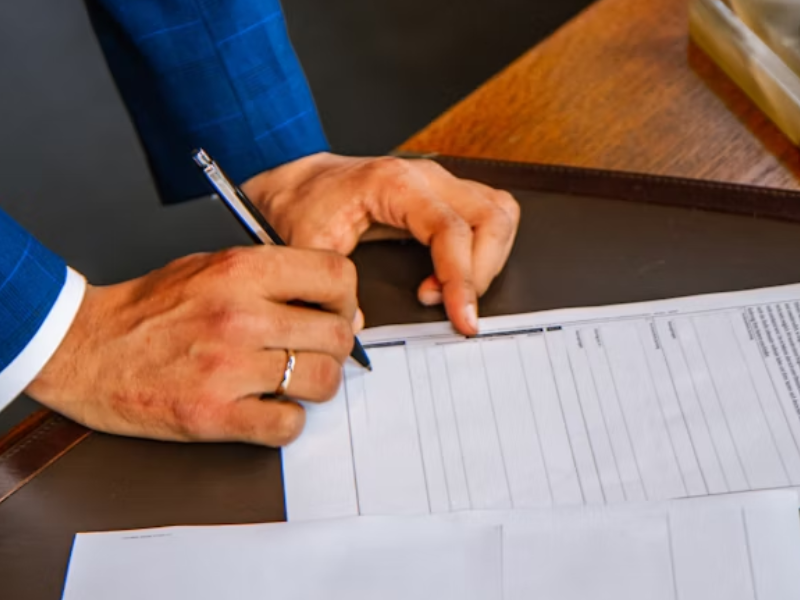 A imagem mostra um homem assinando um documento oficial sobre uma mesa de madeira escura.