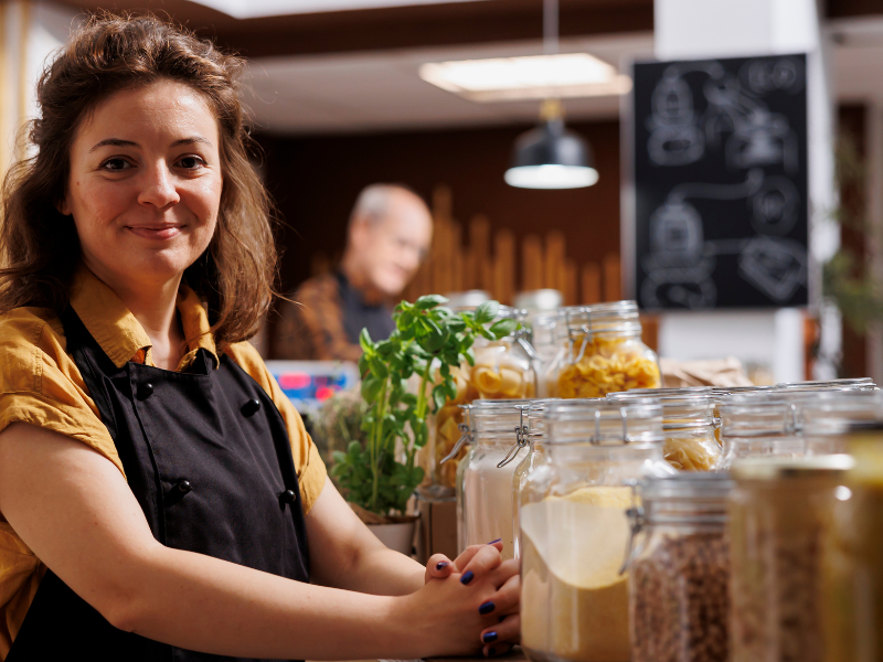 A imagem mostra uma mulher empreendedora em um ambiente que parece ser uma mercearia, loja de produtos naturais ou empório, sugerindo um pequeno negócio próprio, como uma microempresa.