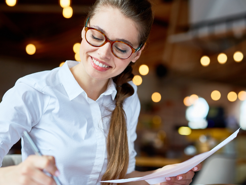 A imagem mostra uma mulher jovem em um ambiente interno, provavelmente um café moderno ou espaço de coworking, com luzes decorativas ao fundo.