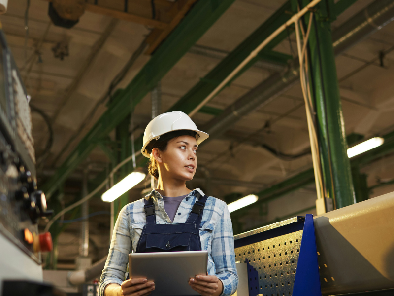 A imagem mostra uma mulher engenheira ou técnica de produção dentro de uma fábrica ou indústria, utilizando capacete branco de segurança e segurando um tablet. Ela está vestindo jardineira jeans com camisa xadrez, sugerindo uma função técnica-operacional.