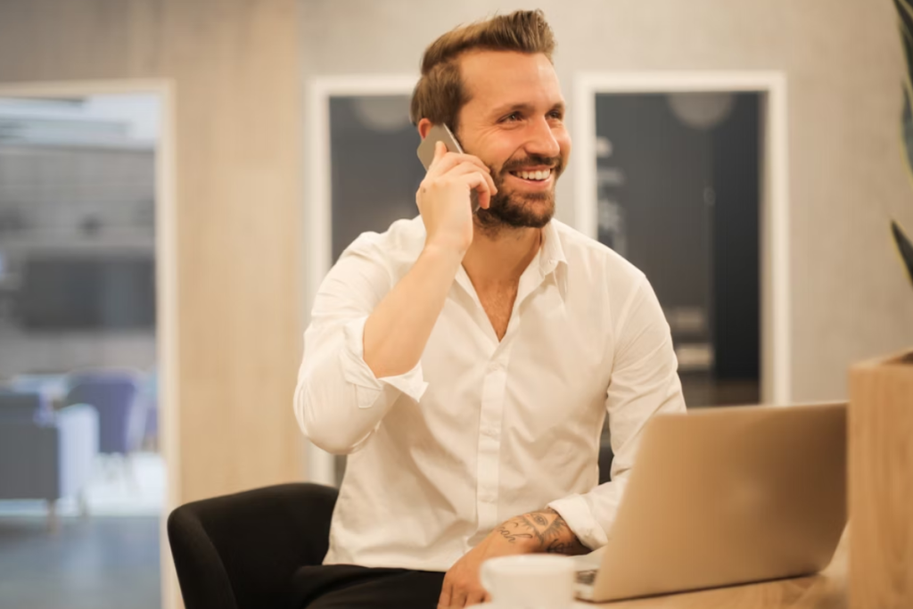A imagem mostra um homem sorridente, com aparência profissional informal, vestindo uma camisa social branca com as mangas dobradas. Ele está sentado em frente a um notebook aberto, enquanto fala ao celular, aparentando estar envolvido em uma conversa agradável ou produtiva.